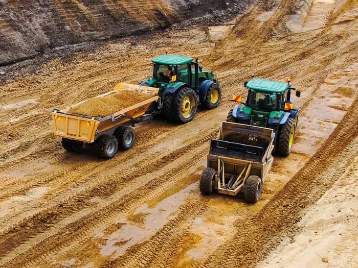 Two green tractors with yellow wheels working in a muddy construction site.